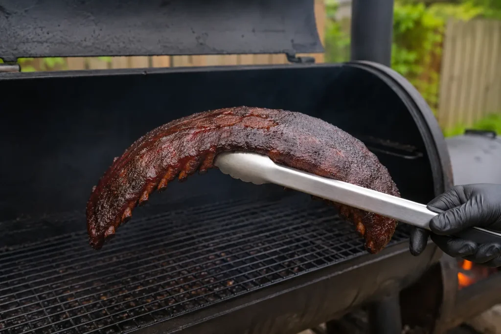 Testing smoked ribs doneness using the bend test with metal tongs showing meat cracking